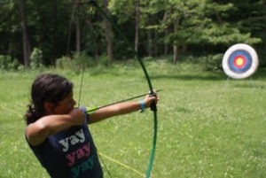 A girl drawing back a bow and arrow aiming at a target.