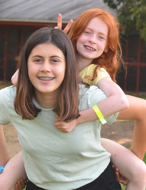 A white teenage girl camper from Camp Kupugani's leadership programs giving another camper a piggyback ride..