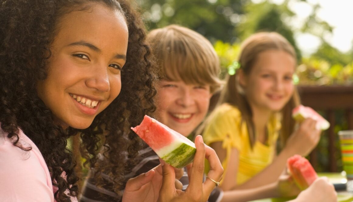 A black girl and white boy and girl building healthy habits by eating a healthy watermelon outside.