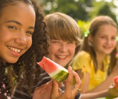A black girl and white boy and girl building healthy habits by eating a healthy watermelon outside.