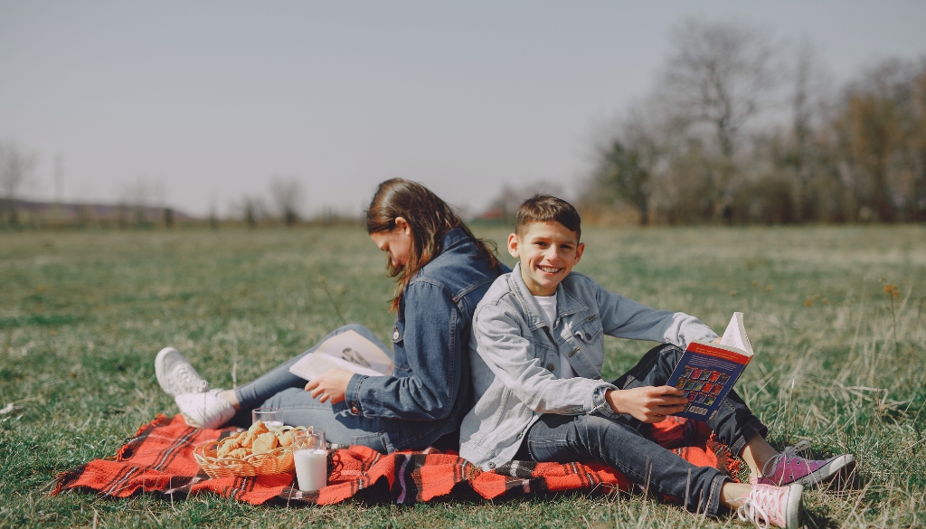 A white boy and girl siblings reading books outdoor alone on a blanket with snacks.