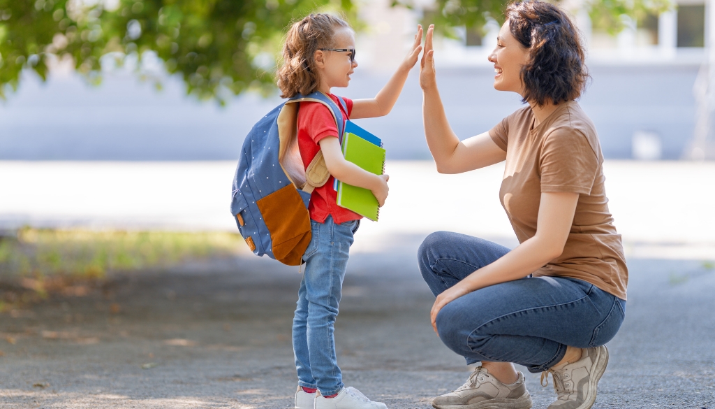 Mom giving daughter high five while standing outside of a school, her daughter is wearing a backpack and holding a folder.